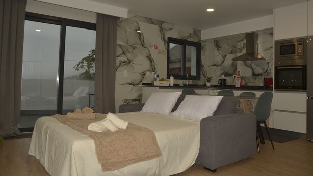 Une chambre d’hôtel moderne dotée d’un grand lit, d’un canapé, d’une kitchenette et offrant une vue sur l’extérieur grâce à une porte coulissante.