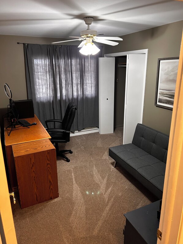 A compact room with a ceiling fan, a desk with a ring light, a black office chair, a gray sofa, and a door leading to another room. - Spacious 3-bedroom house in beautiful Brighton. (Brighton)