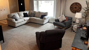 A living room with a sectional sofa, a small armchair, a lamp, and a clock on the wall. - Spacious 3-bedroom house in beautiful Brighton. (Brighton)