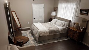 A bedroom with a bed, a chair, a dresser, a lamp, a door, and a painting on the wall. - Spacious 3-bedroom house in beautiful Brighton. (Brighton)