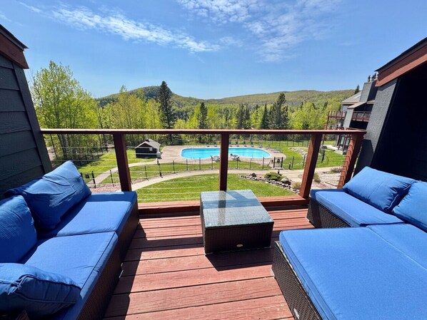 A deck with blue cushioned seating and a glass-top table, overlooking a swimming pool and a green landscape.