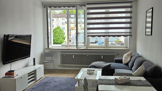 A modern living room with a flat-screen TV, sofa, coffee table, and a view of buildings through large windows.