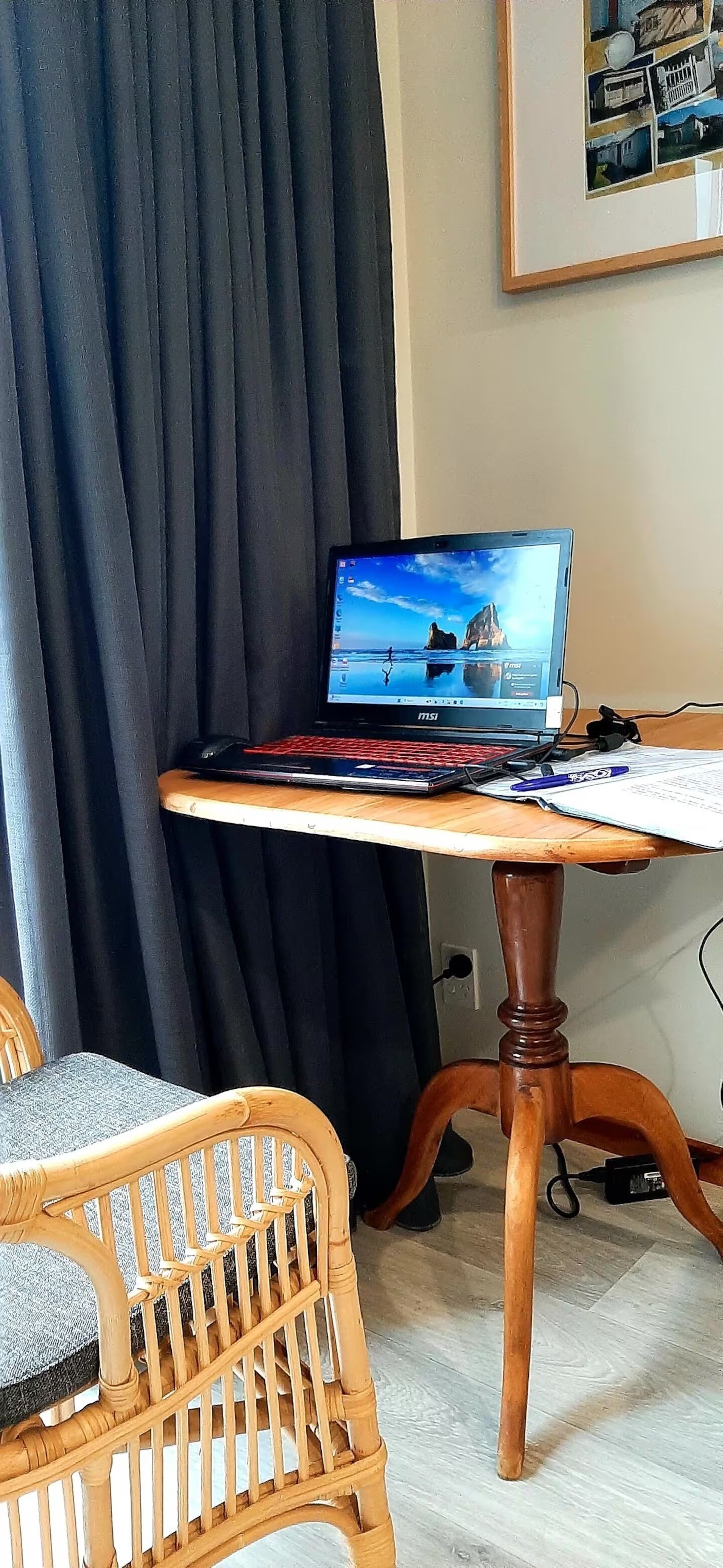 Un portatile su un tavolo di legno con vista mare sullo schermo, una sedia in vimini e un quadro appeso al muro.