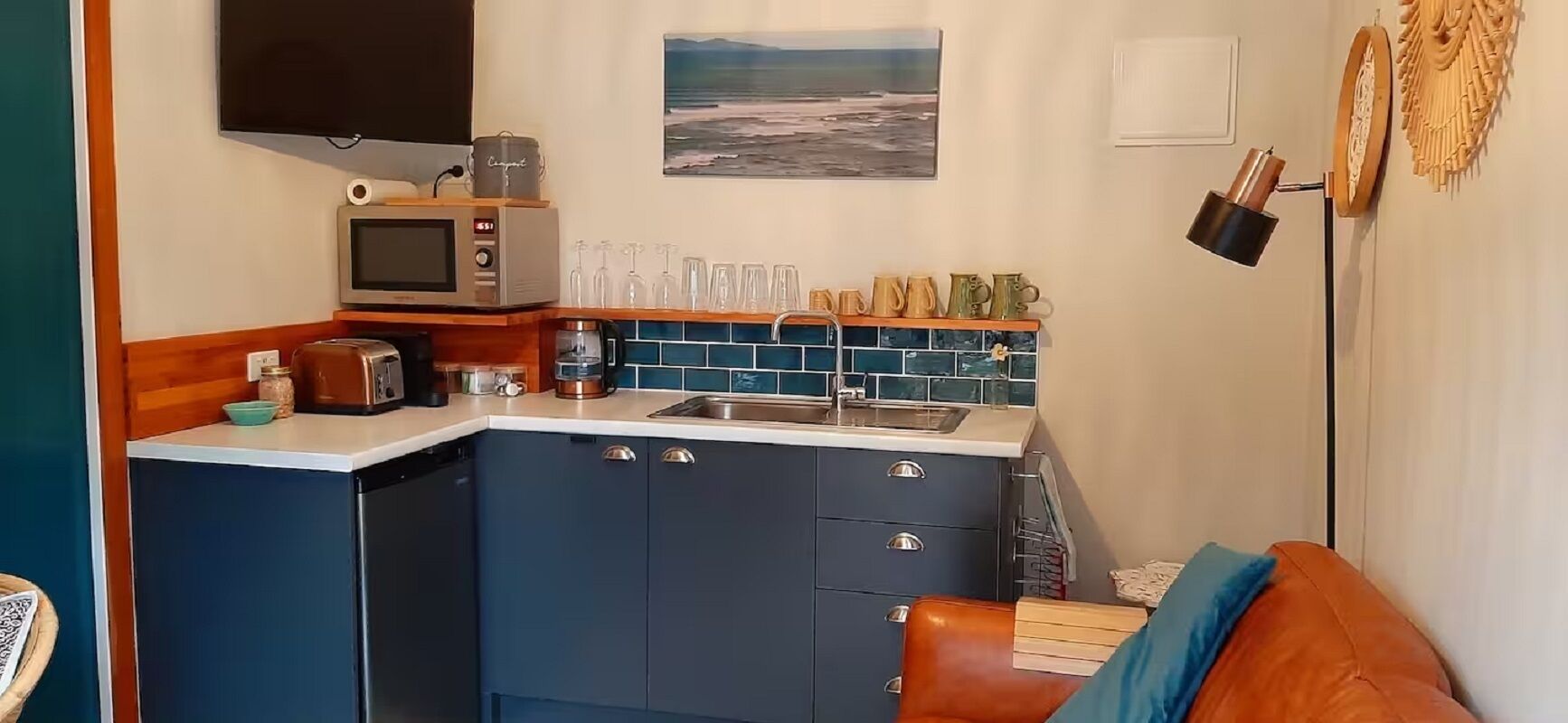 Cocina compacta con microondas, tostadora y cafetera. Hay un lavabo con salpicadero azul y una ventana pequeña. Se ve un sofá de cuero marrón al frente.