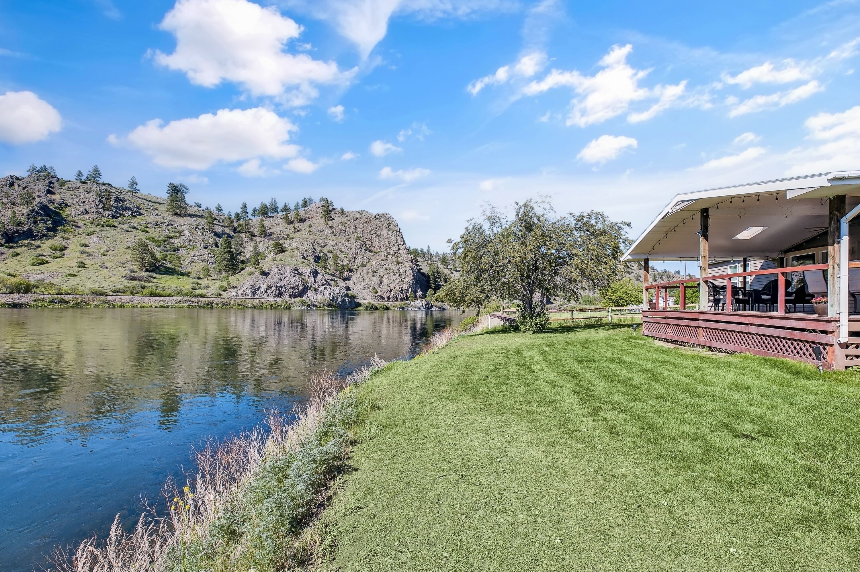 A lakeside property with a grassy lawn, a wooden deck, and a view of a rocky hill.