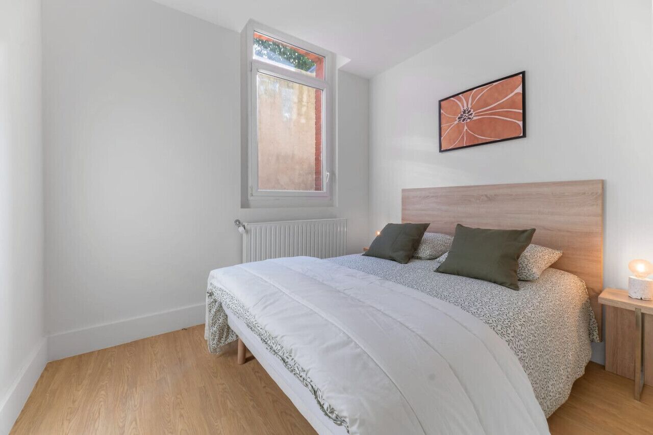 Un dormitorio con una cama grande, una ventana, un radiador, una mesita de noche con una lámpara y un cuadro enmarcado en la pared.
