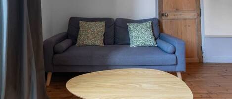 A room with a grey sofa, two patterned cushions, a wooden door, and a round wooden table.
