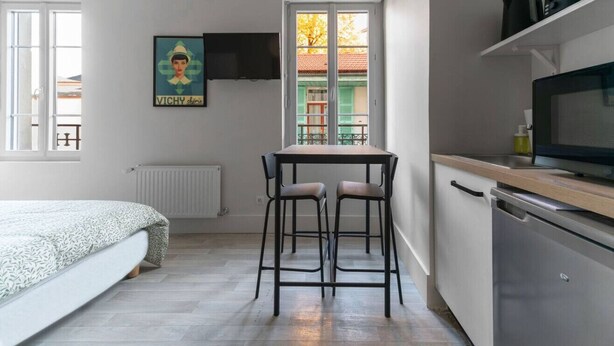 A compact room with a bed, a small table with two chairs, a TV mounted on the wall, and a kitchen area with a dishwasher and a microwave. - Le Desroziers 312 - 31 avenue Victoria - Vichy (Vichy)