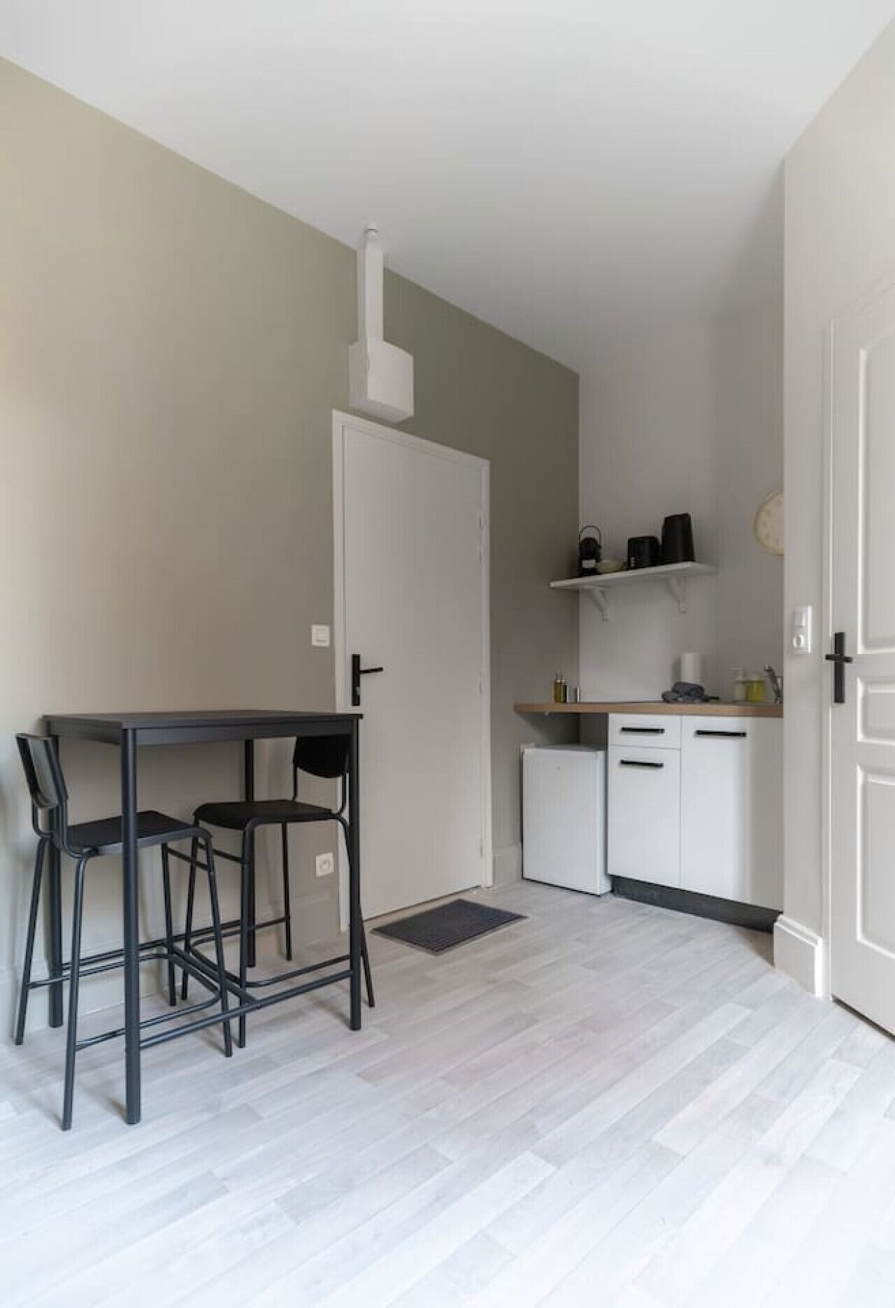 Habitació petita i neta amb una porta blanca, una cuina americana, una taula amb dues cadires i un terraig blanc.