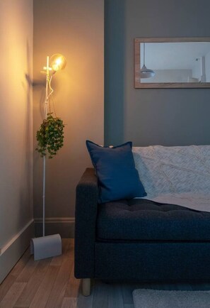 Ein modernes Wohnzimmer mit einem dunkelgrauen Sofa, einem blauen Kissen, einer Stehlampe mit hängender Pflanze und einem gerahmten Bild an der Wand.