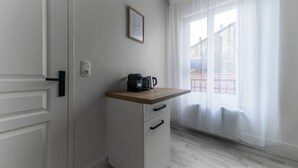 Una pequeña kitchenette con una cafetera sobre una encimera de madera, un cuadro en la pared y vistas a un edificio a través de cortinas translúcidas.