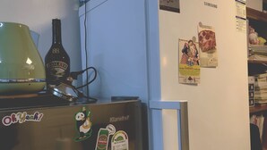 Et køkken med et køleskab, en mikrobølgeovn og en elkedel. Der er magneter og beskeder på køleskabet, og en flaske Bailey's øl står på bordet.