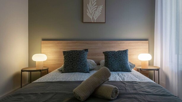 Un lit bien fait, recouvert d’une couvre-lit à motifs, avec deux oreillers et une serviette enroulée. Deux lampes de chevet sont placées de chaque côté du lit.