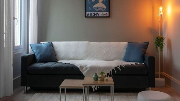 A living room with a dark sofa, a small coffee table, and a round ottoman. There is a framed picture on the wall and a floor lamp.