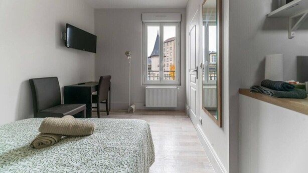 A bedroom with a bed, a desk, a chair, a TV, a window, and a view of buildings outside.