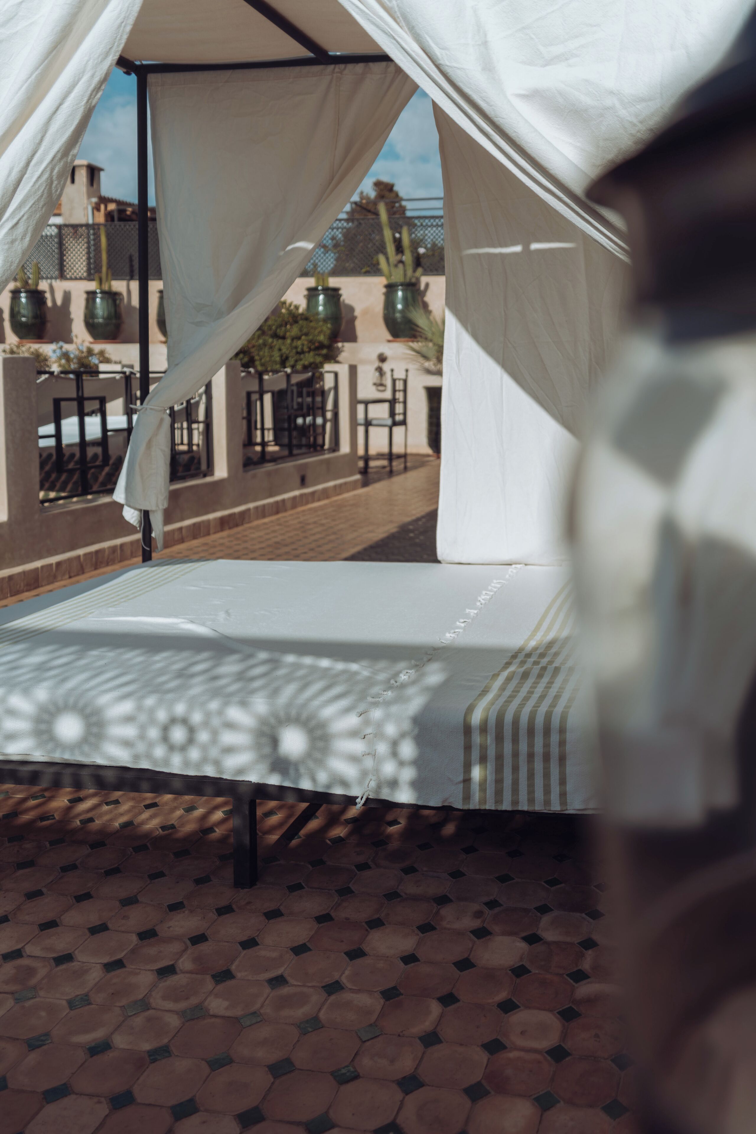 A bed with a canopy, a tiled floor, and a view of buildings and plants in the background.