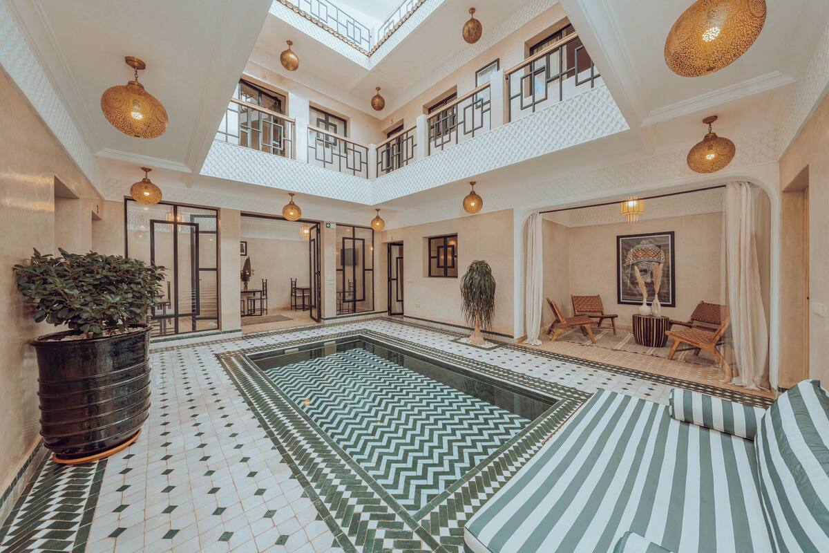 A spacious indoor pool area with a geometric tile floor, multiple seating areas, and a high ceiling with hanging lights.