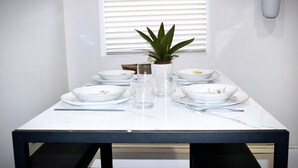 A dining table set with plates, glasses, and cutlery, a potted plant, and a window with blinds.