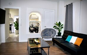 A living room with a black sofa, a glass coffee table, a grey armchair, and a potted plant. - King Bed / Coffee Bar / Meditation Room / Remote Work / PacMan / 30min to NYC (Elizabeth)