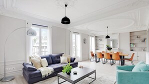 En moderne stue med en mørkeblå sofa, et sofabord og turkise lænestole. Der er et spiseområde med et træbord og orange stole.