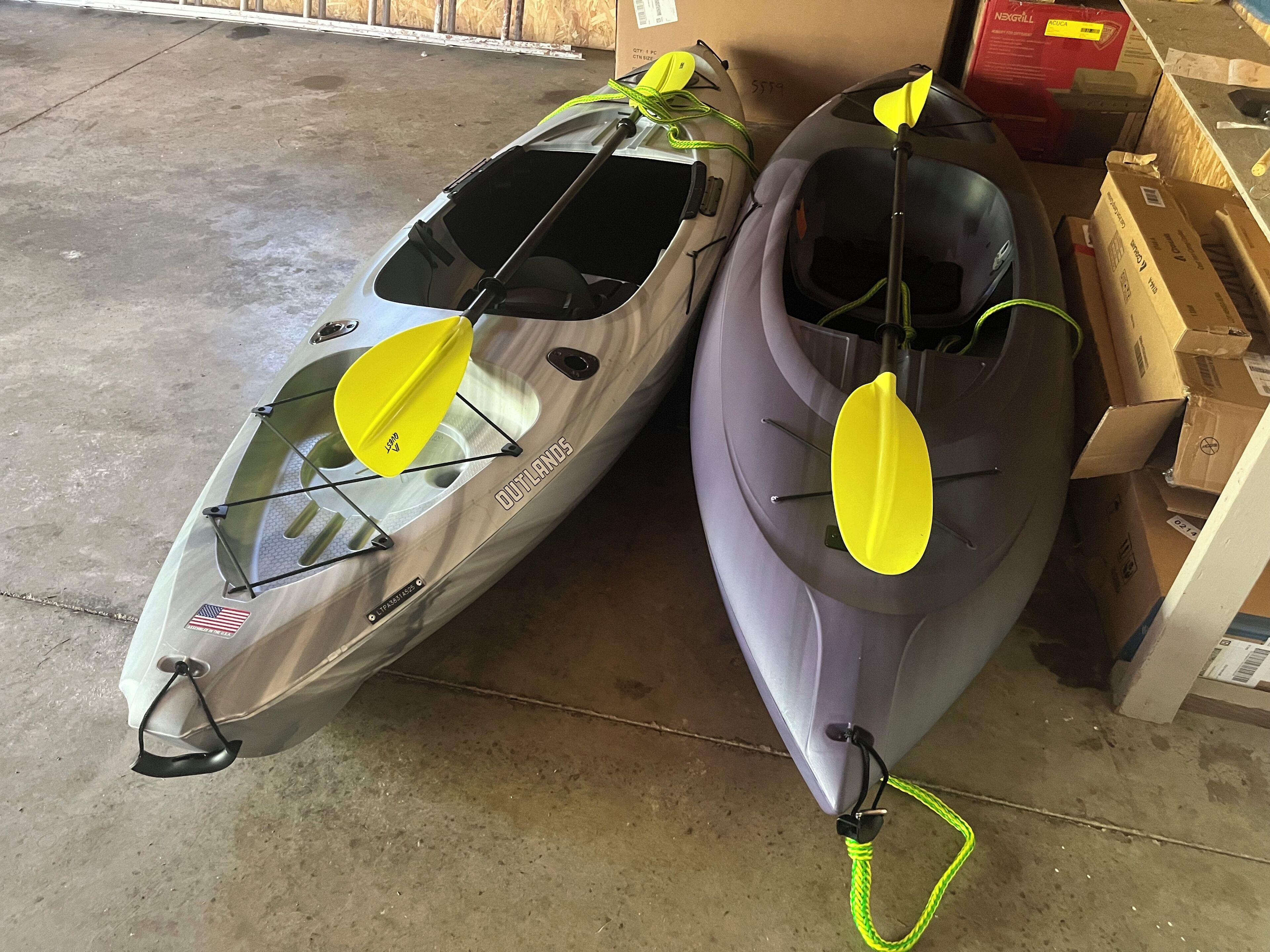 Two kayaks with yellow paddles, one silver and one gray, in a garage with cardboard boxes and a wooden wall in the background.