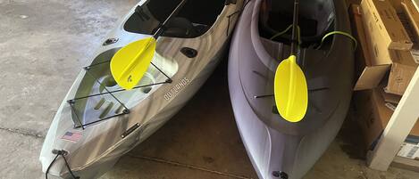 Two kayaks with yellow paddles, one silver and one gray, in a garage with cardboard boxes and a wooden wall in the background.