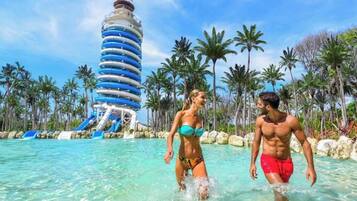 Área de piscina en un resort con agua cristalina, palmeras y una estructura de tobogán distintiva.