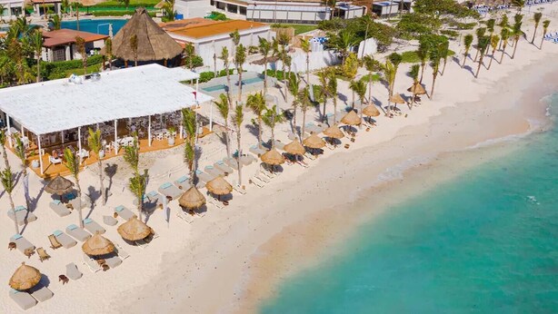 Vista aérea de un resort en la playa, con filas de tumbonas y sombrillas de paja, una piscina y un edificio blanco con techo de paja.
