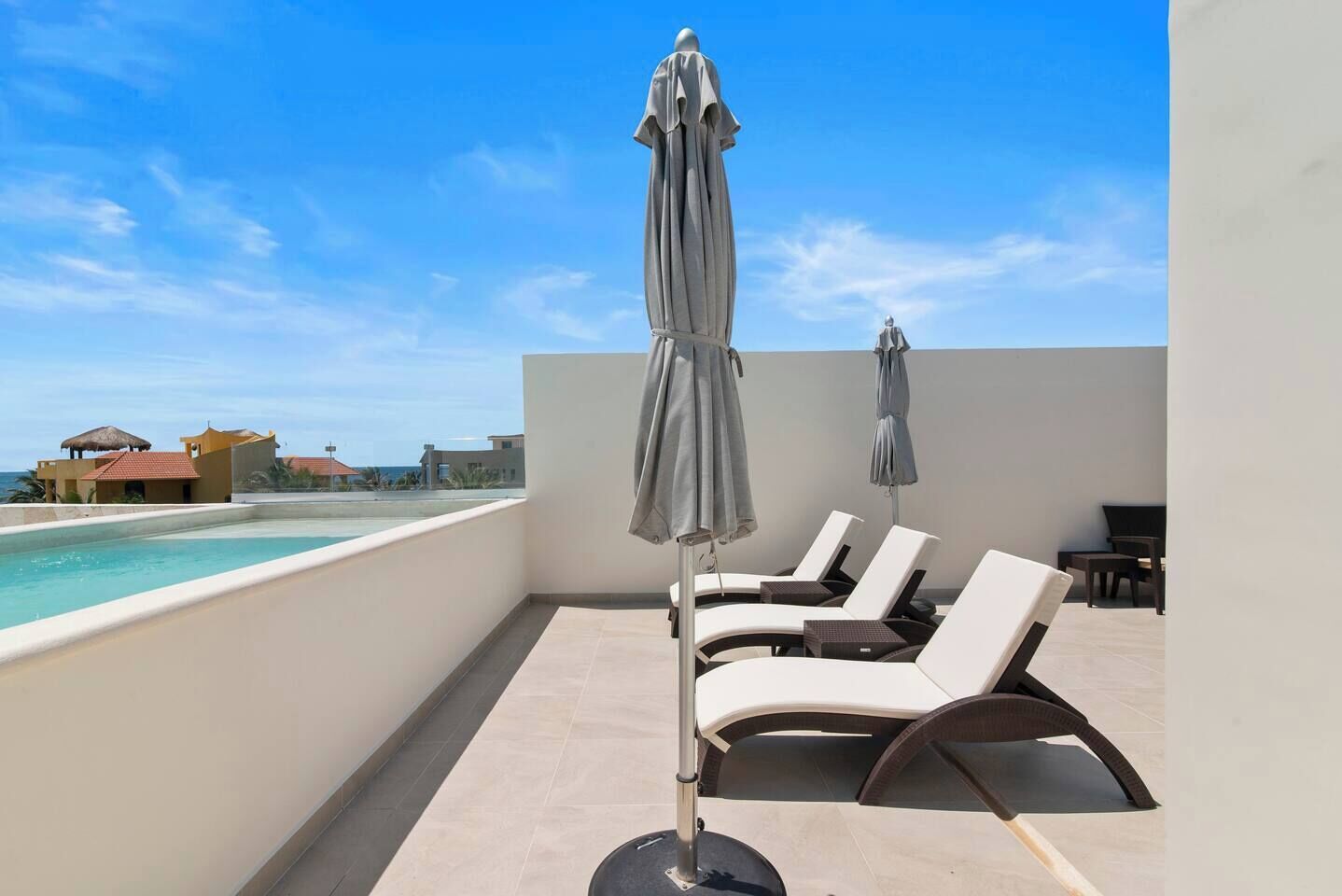 Un espace piscine aménagé sur le toit, avec des chaises longues et des parasols, offrant une vue dégagée sur un ciel bleu et des bâtiments éloignés.