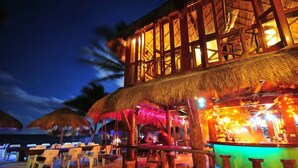 Un hotel frente a la playa con zona de bar, mesas al aire libre y sombrillas de paja.