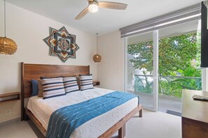 Ein Schlafzimmer mit einem großen Bett, einem Deckenventilator, einem dekorativen Wandbild und Blick auf Grün hinter einer Schiebetür.