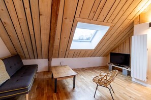 A cozy attic room with a sloped ceiling, a wooden floor, a sofa, a table, a wicker chair, and a TV. - Le Repaire Du Nandrion - For Two (TREVIGNIN)