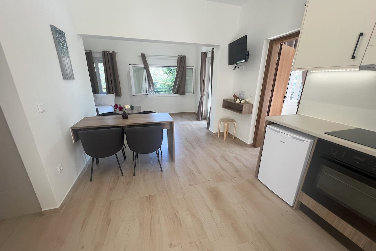 A modern kitchen with a dining area, wooden flooring, a small refrigerator, and a TV mounted on the wall.