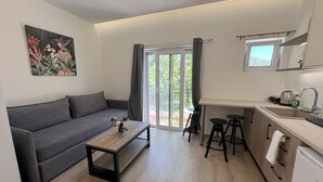 Un soggiorno moderno con un divano grigio, un tavolino in legno e un quadro con fiori. Accostata si trova una cucina con un lavello, un bollitore e un frigorifero.