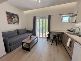 Una sala de estar moderna con un sofá gris, una mesa de centro de madera y un cuadro de flores. Junto a ella, una cocina con un fregadero, una tetera y un refrigerador.