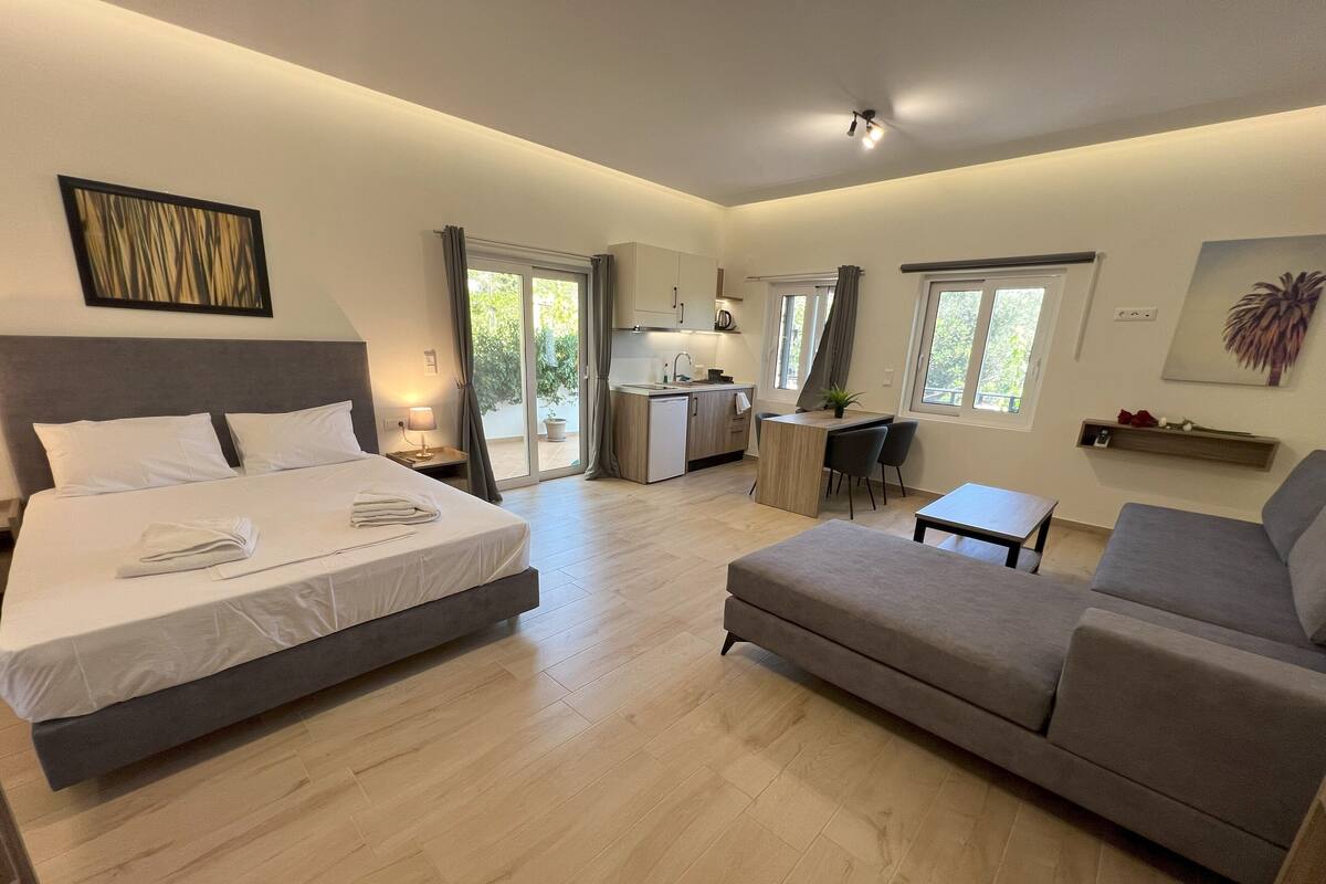 A modern hotel room with a large bed, a sofa, a desk, and a kitchenette.
