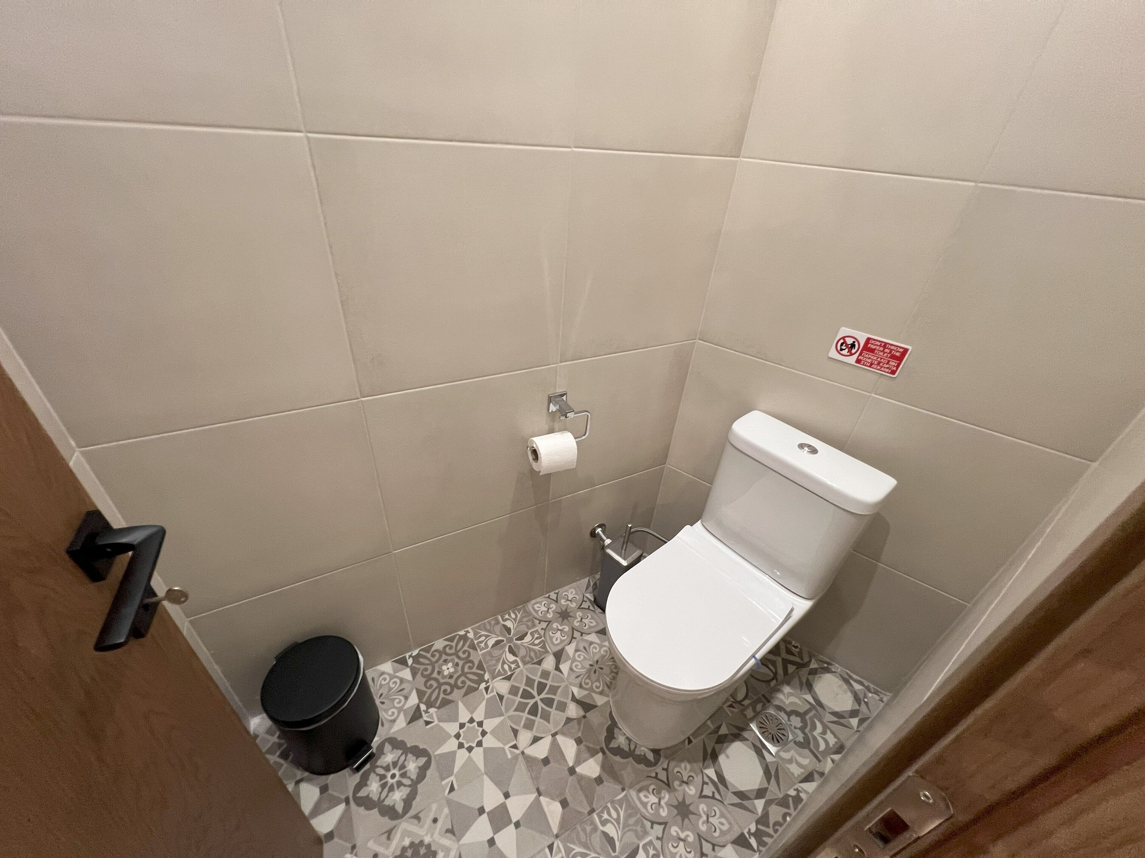 A small, tiled bathroom with a toilet, a trash bin, and a paper towel dispenser.