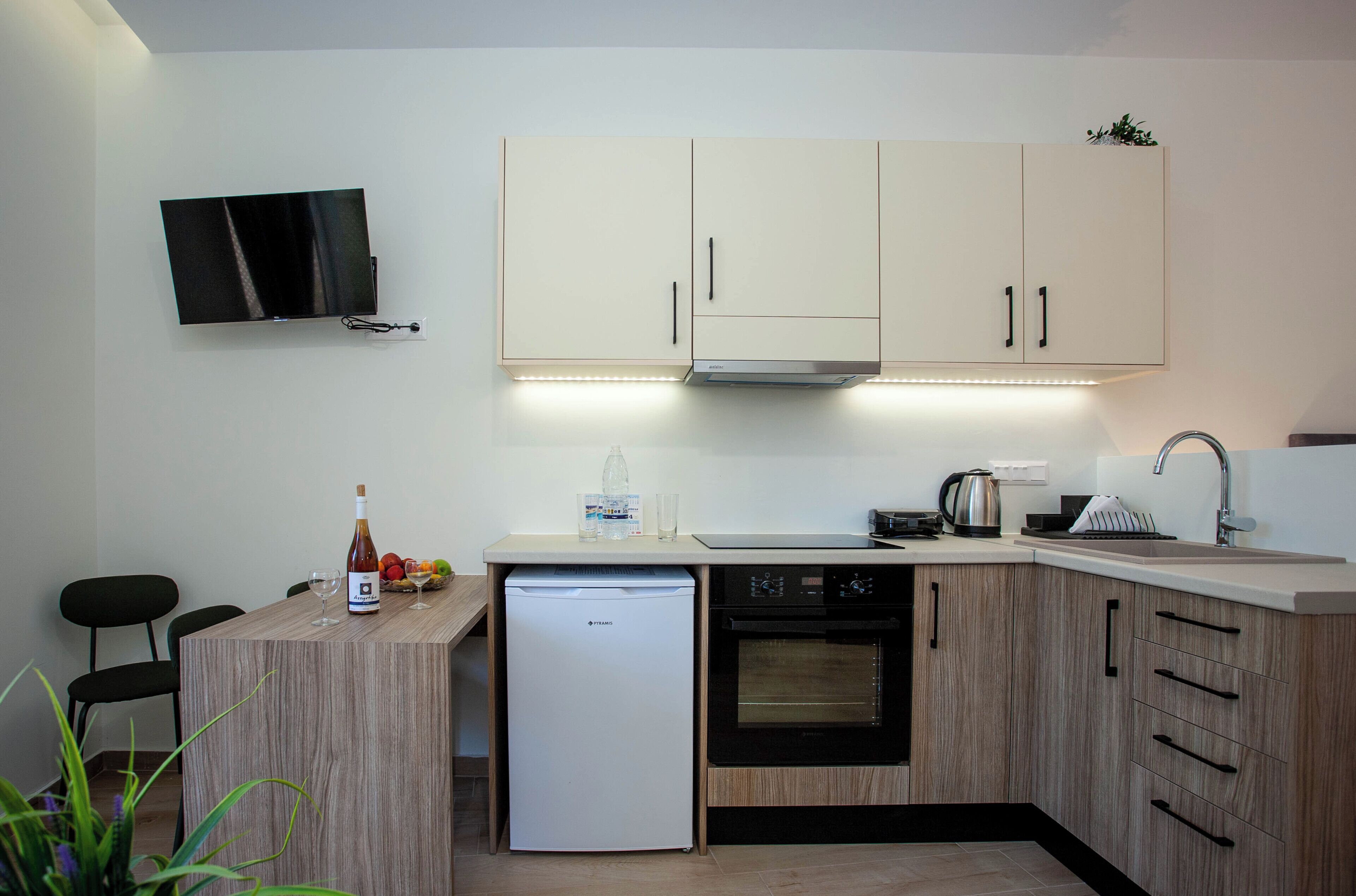 A compact kitchen with a built-in oven, a dishwasher, a small refrigerator, and a flat-screen TV mounted on the wall.