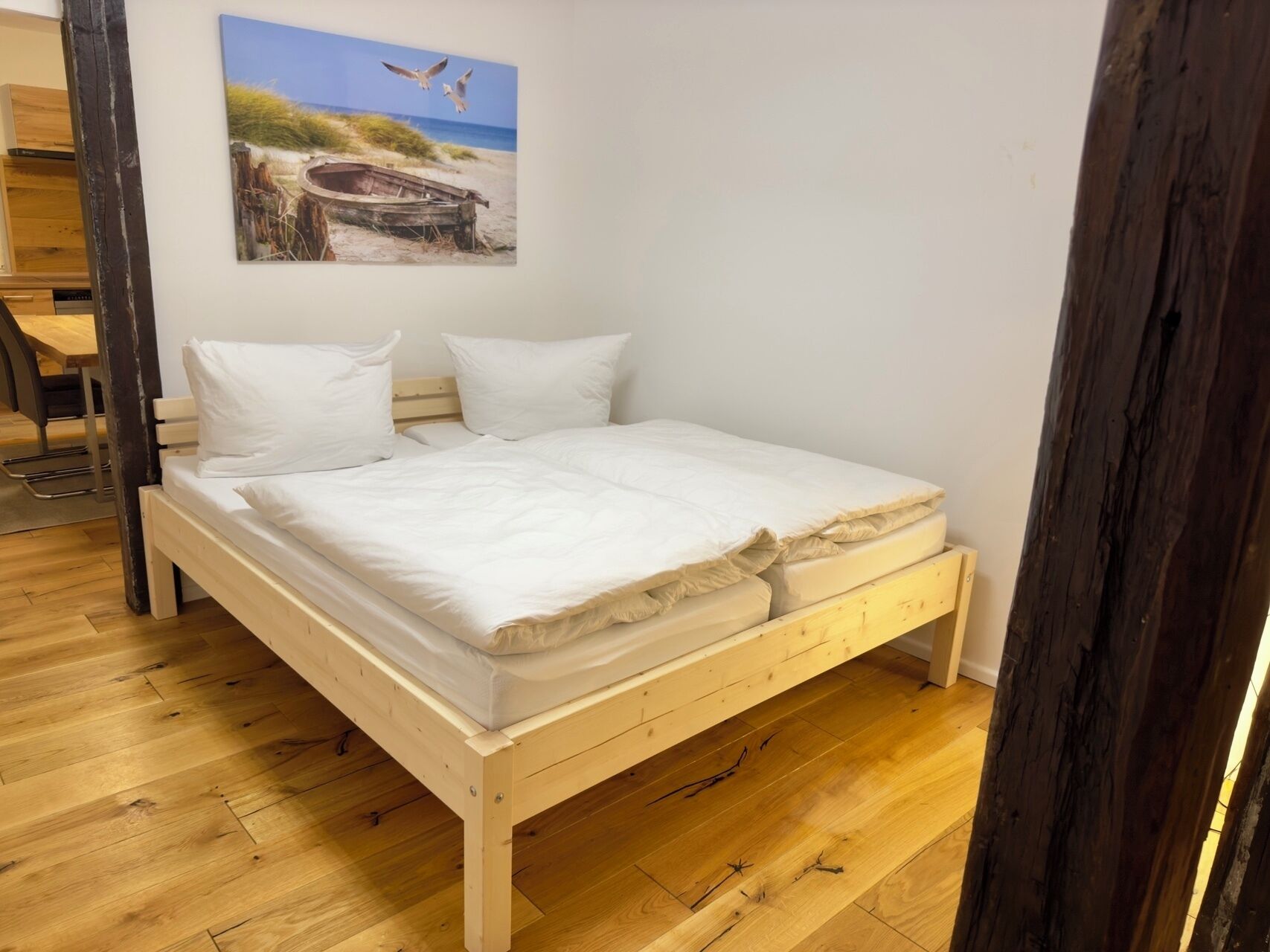 A wooden bed with white bedding in a room with wooden flooring and a beach-themed painting on the wall.