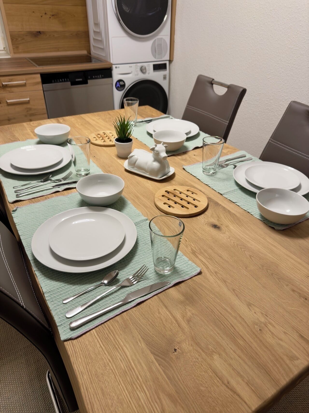 Ein Esstisch ist für vier Personen gedeckt, mit Tellern, Besteck und Gläsern. Auf dem Tisch stehen ein kleiner Topfpflanze und eine Zierfigur eines Keramikziegens.
