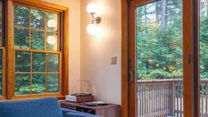 A room with a wooden deck, a blue sofa, a wooden desk with a record player, and a large window with a view of trees.