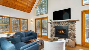 A spacious living room with a stone fireplace, large windows, and a television mounted above it.