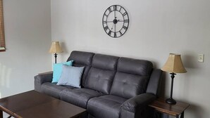 Een woonkamer met een donker leren bank, twee bijzettafels met lampen, een houten salontafel en een wandklok.