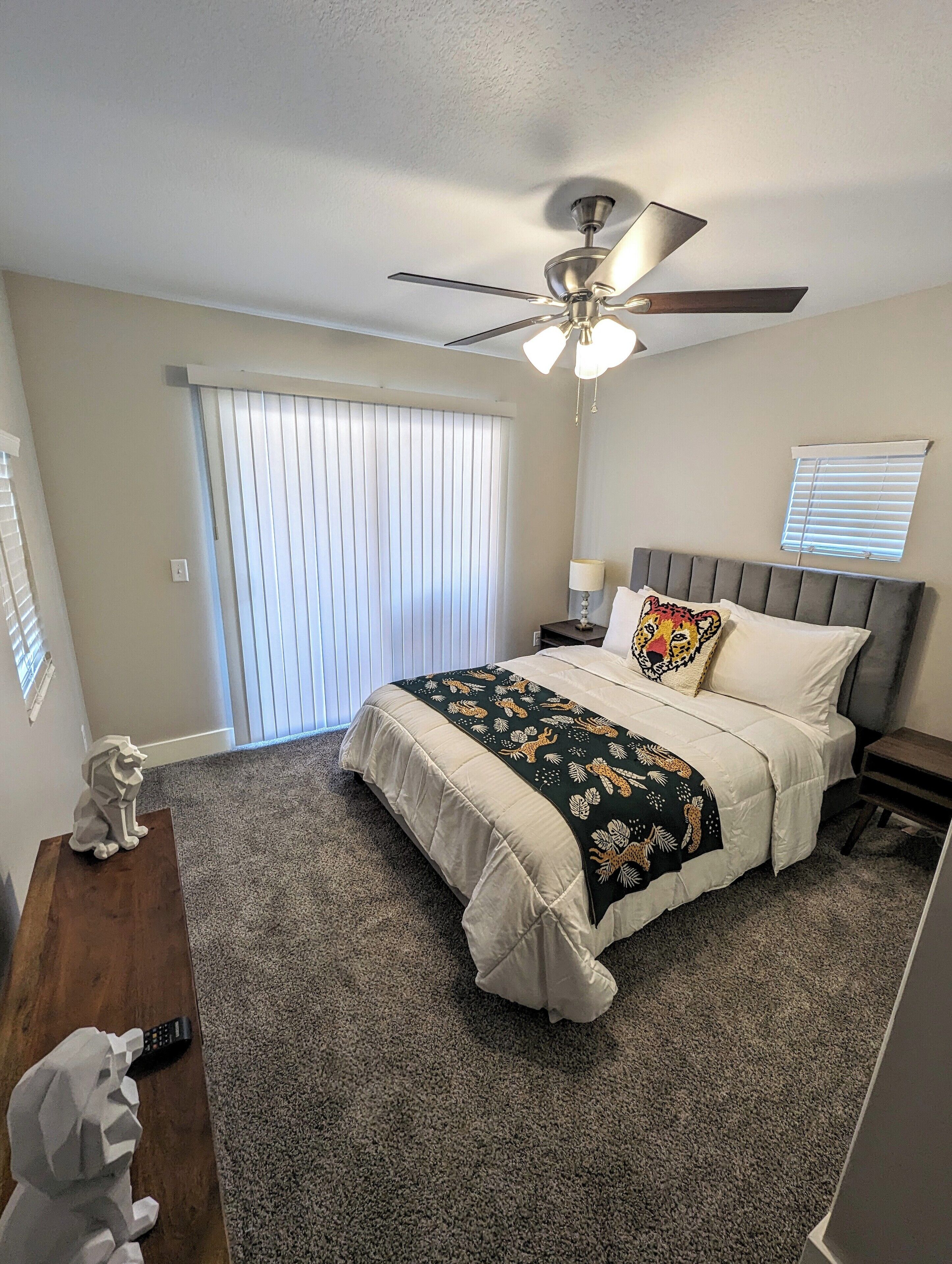 Une chambre à coucher comprenant un lit, un ventilateur de plafond, une table de chevet, une lampe, une fenêtre avec des stores et une porte.