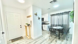 Una cocina moderna con armarios blancos, un comedor con mesa y sillas, y una puerta con un pequeño estante que sostiene una planta y una taza.