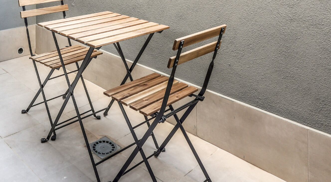 Una mesa de madera plegable y dos sillas con estructura de metal, ubicadas sobre un piso de baldosas frente a una pared con textura.