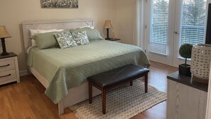 Quarto com cama, mesas de cabeceira, um banco, porta com persianas e um quadro na parede.
