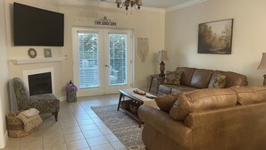 Uma sala de estar com um sofá de couro marrom, uma poltrona estampada, lareira, televisão e ventilador de teto.