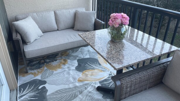 Uma varanda com um sofá cinza, uma mesa de vidro com um vaso de flores e um chão estampado.