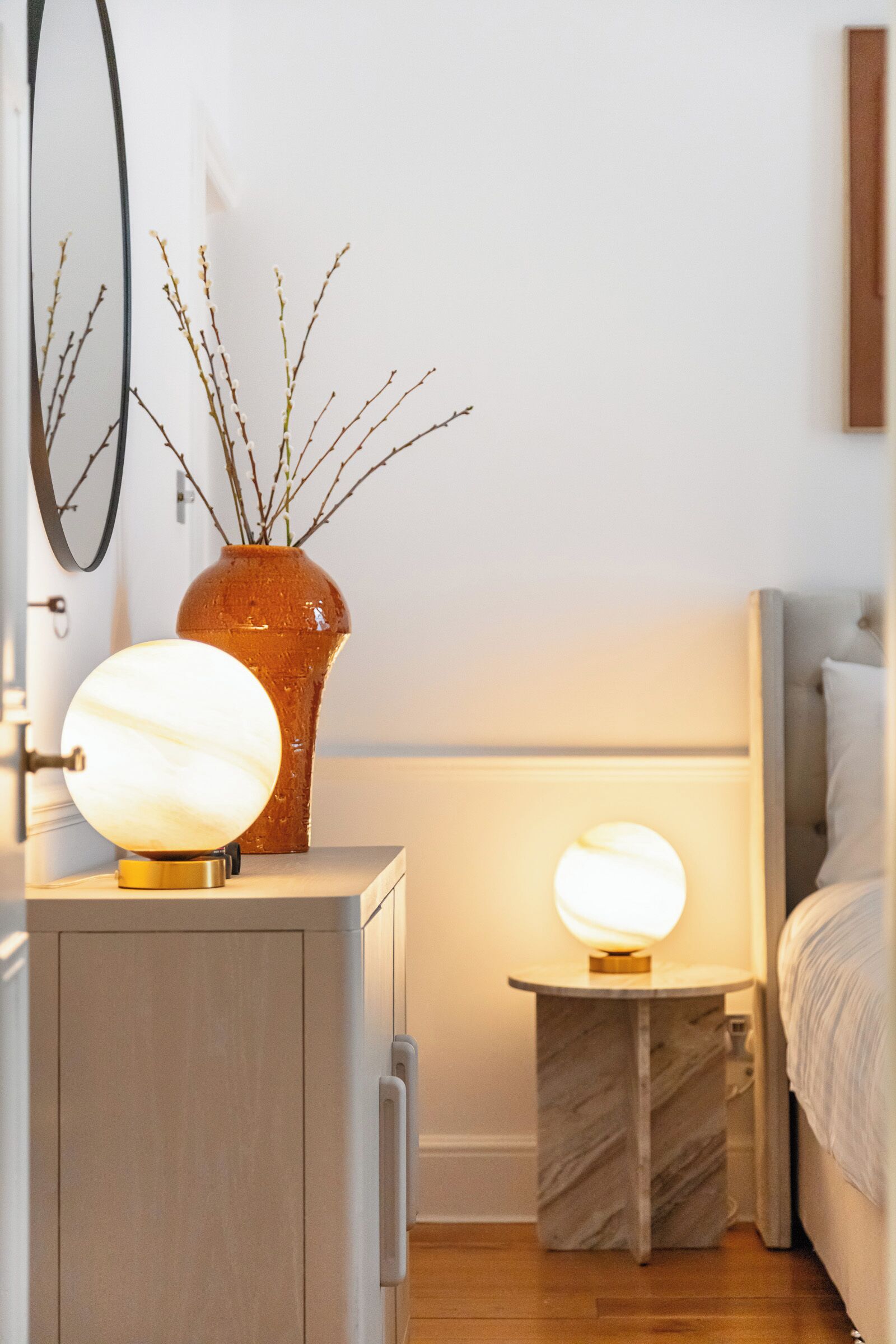 A bedroom with a bed, bedside table, lamp, and a vase with branches.